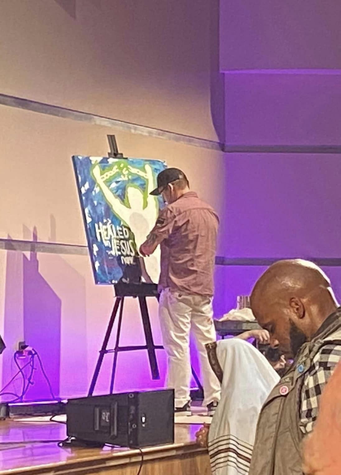 Jim McMahel painting a figure with raised arms and the words Healed by Jesus on a church stage under purple light