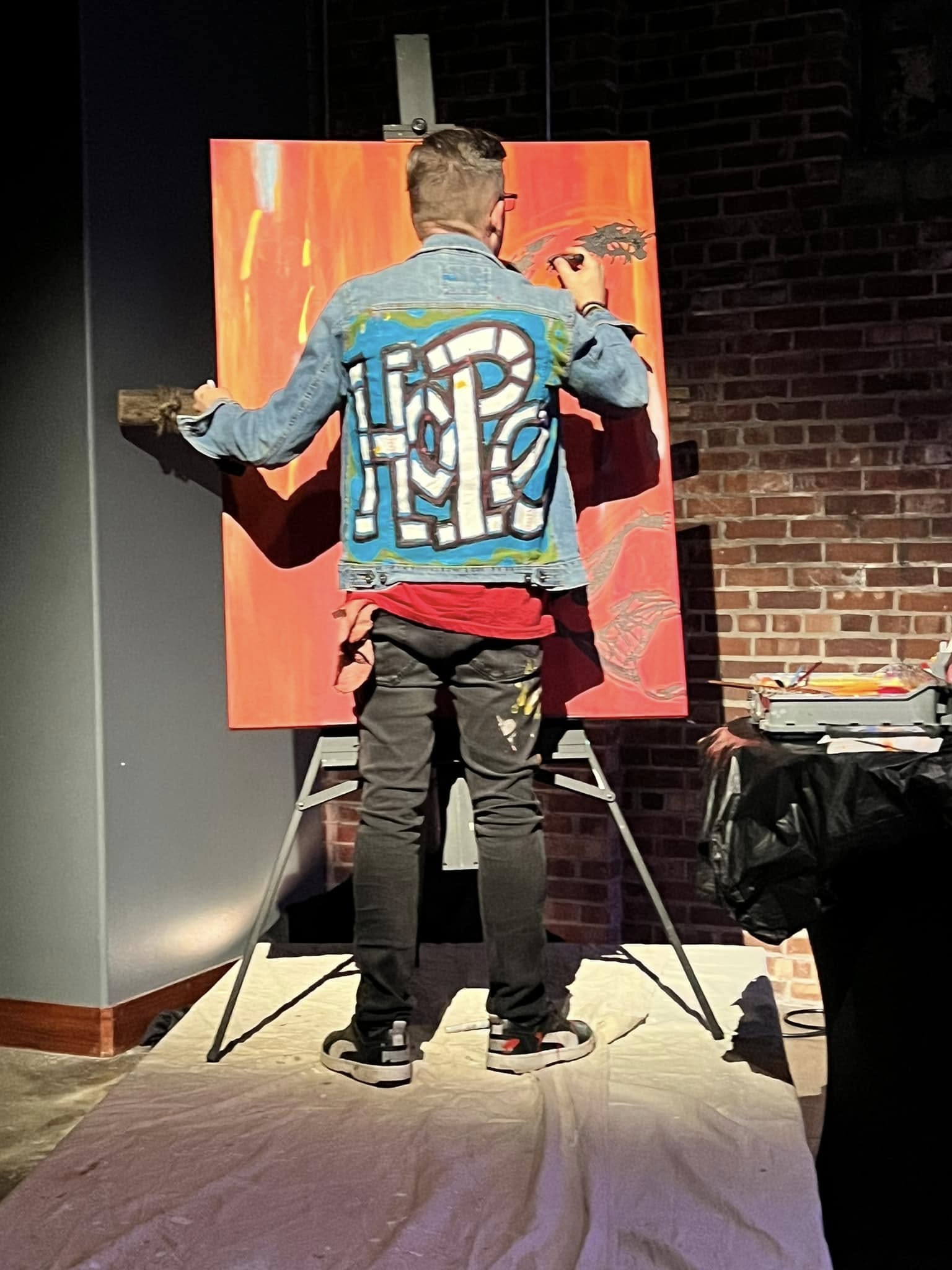 Jim McMahel seen from behind in a denim jacket lettered HOPE, working on an orange canvas